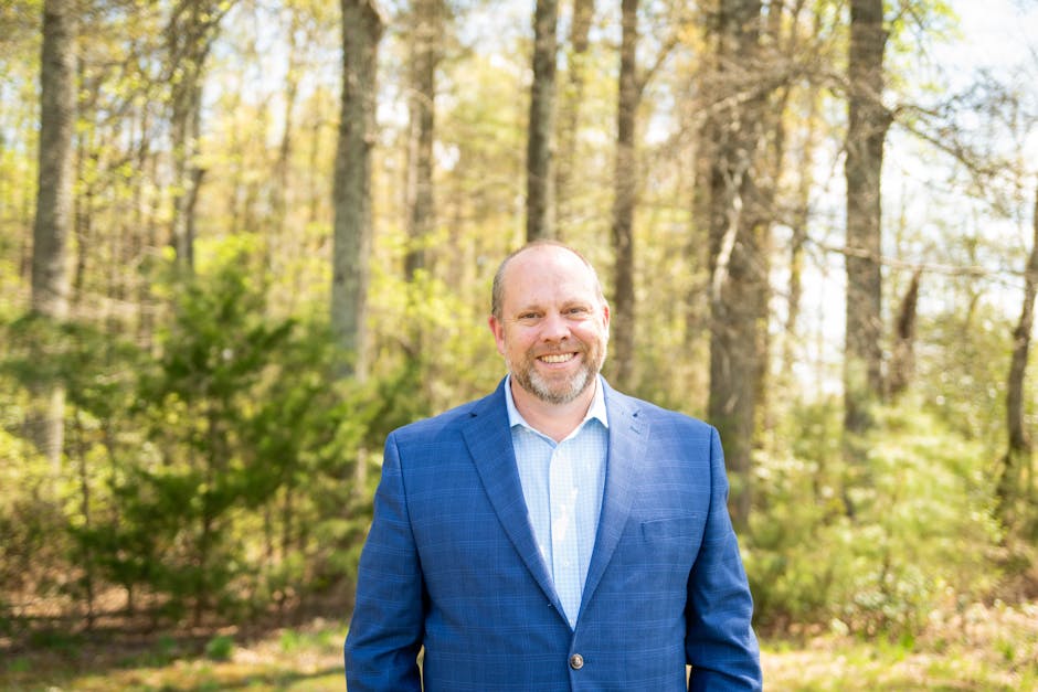 Middle-aged man smiling in outdoor forest with a suit jacket, trees in background.