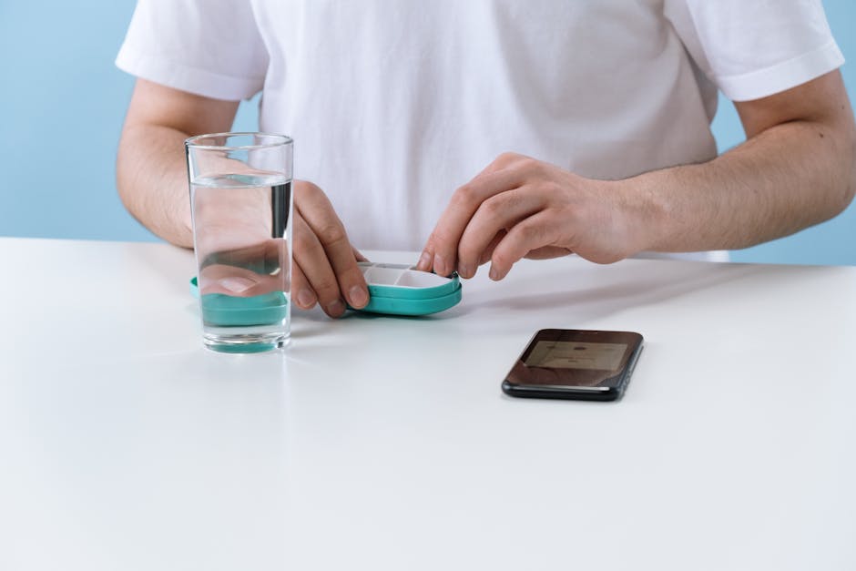 A person arranging pills with a weekly organizer, next to a glass of water and a smartphone.