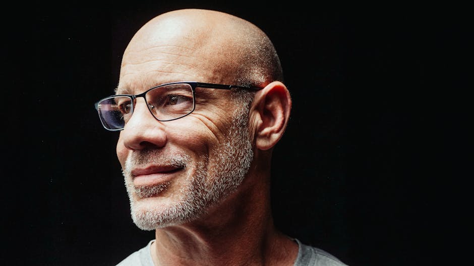 Close-up portrait of a bald man with eyeglasses, smiling against a dark background.