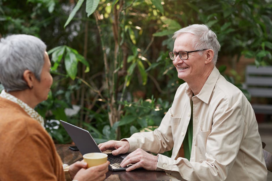 Happy senior couple sitting in a garden cafe, enjoying coffee and conversation with a laptop.