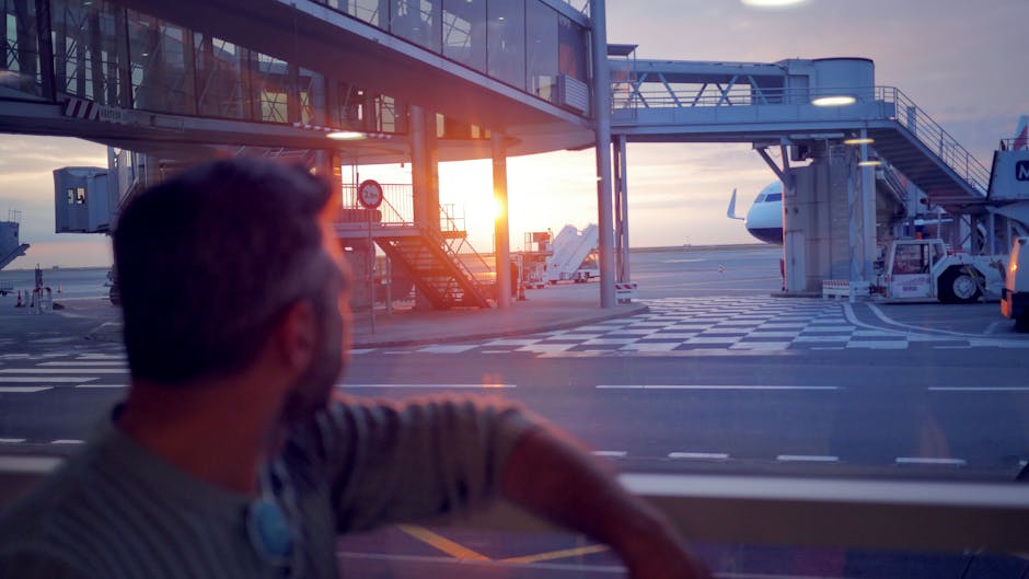 A thoughtful man gazes at the sunset from an airport terminal window, with planes in view.
