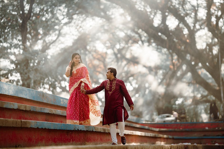 A couple in traditional attire walking hand in hand outdoors with sunlight filtering through trees.