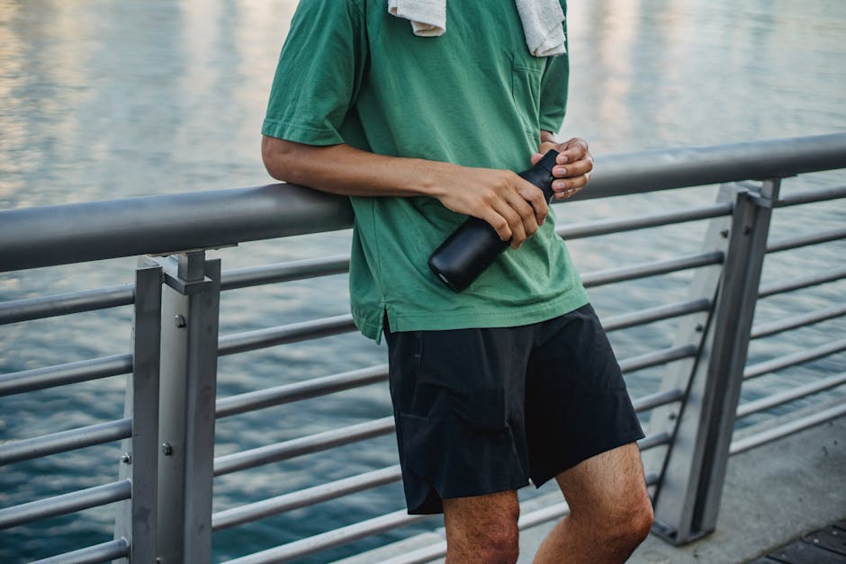 Male in activewear leaning on a railing holding a water jug by the water, post-exercise.