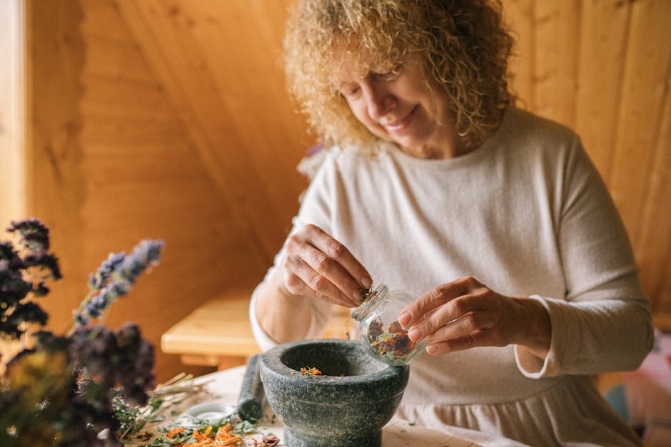 Mature woman in white dress grinding dried flowers using a mortar and pestle indoors.