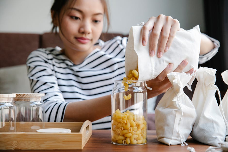 Crop ethnic female adding uncooked pasta from package into transparent shiny jar on wooden table
