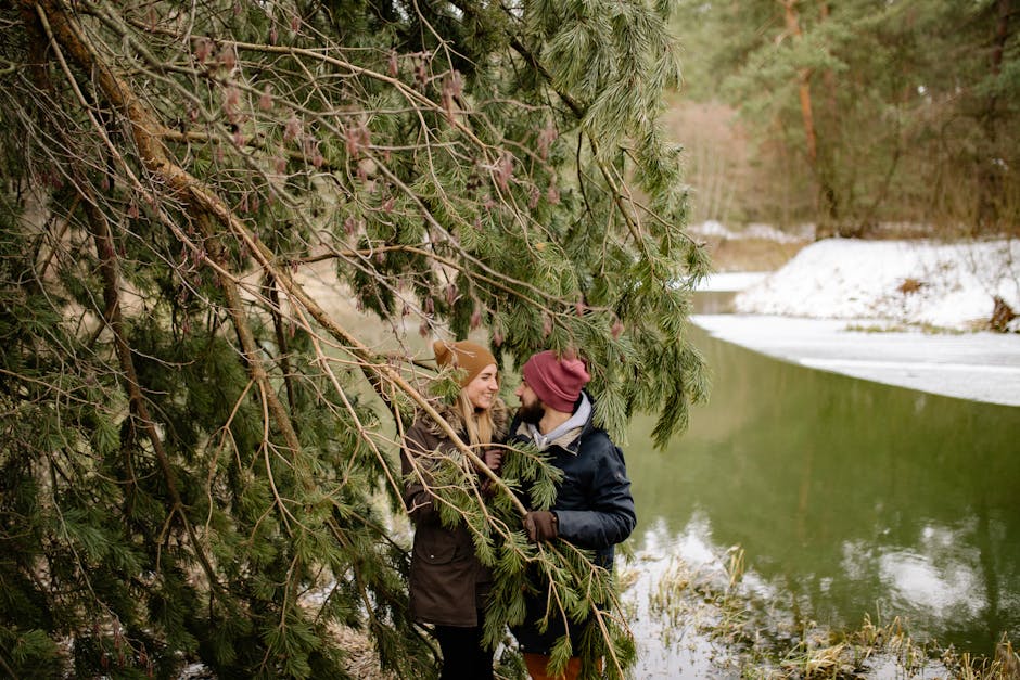 Couple smiling near a snowy river with pine trees during winter. Love and warmth in nature.