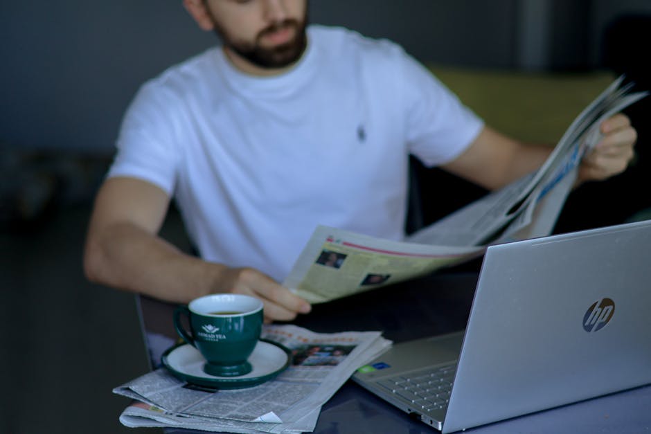A man in a white t-shirt reads a newspaper, with a laptop and coffee nearby. Indoor setting.