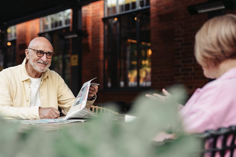 A cheerful elderly couple sitting outdoors, engaging in conversation and reading a newspaper.