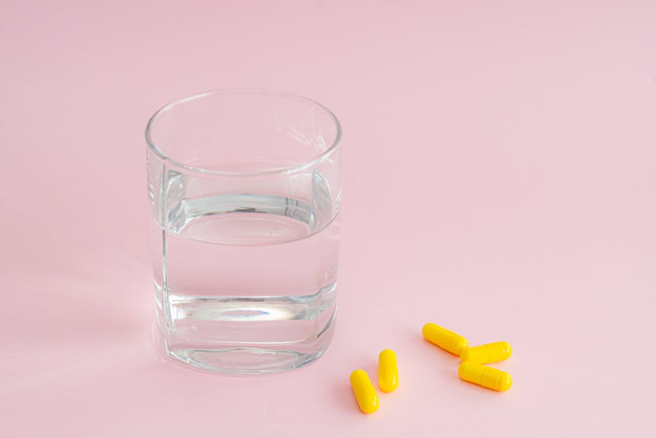 A clear glass of water next to yellow capsules on a pink background, symbolizing health care.