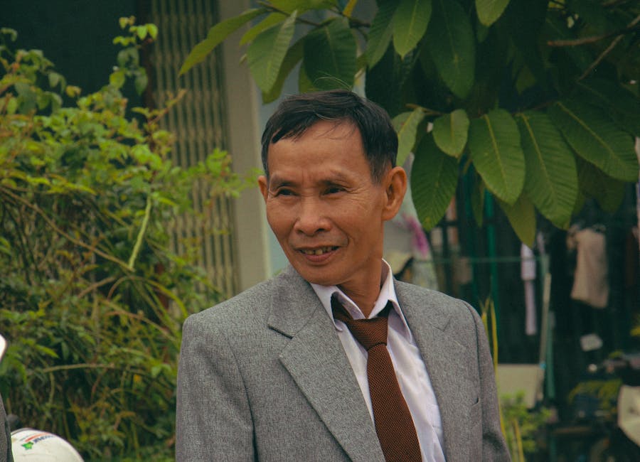 Smiling man in a gray suit and tie standing outdoors with greenery in the background.
